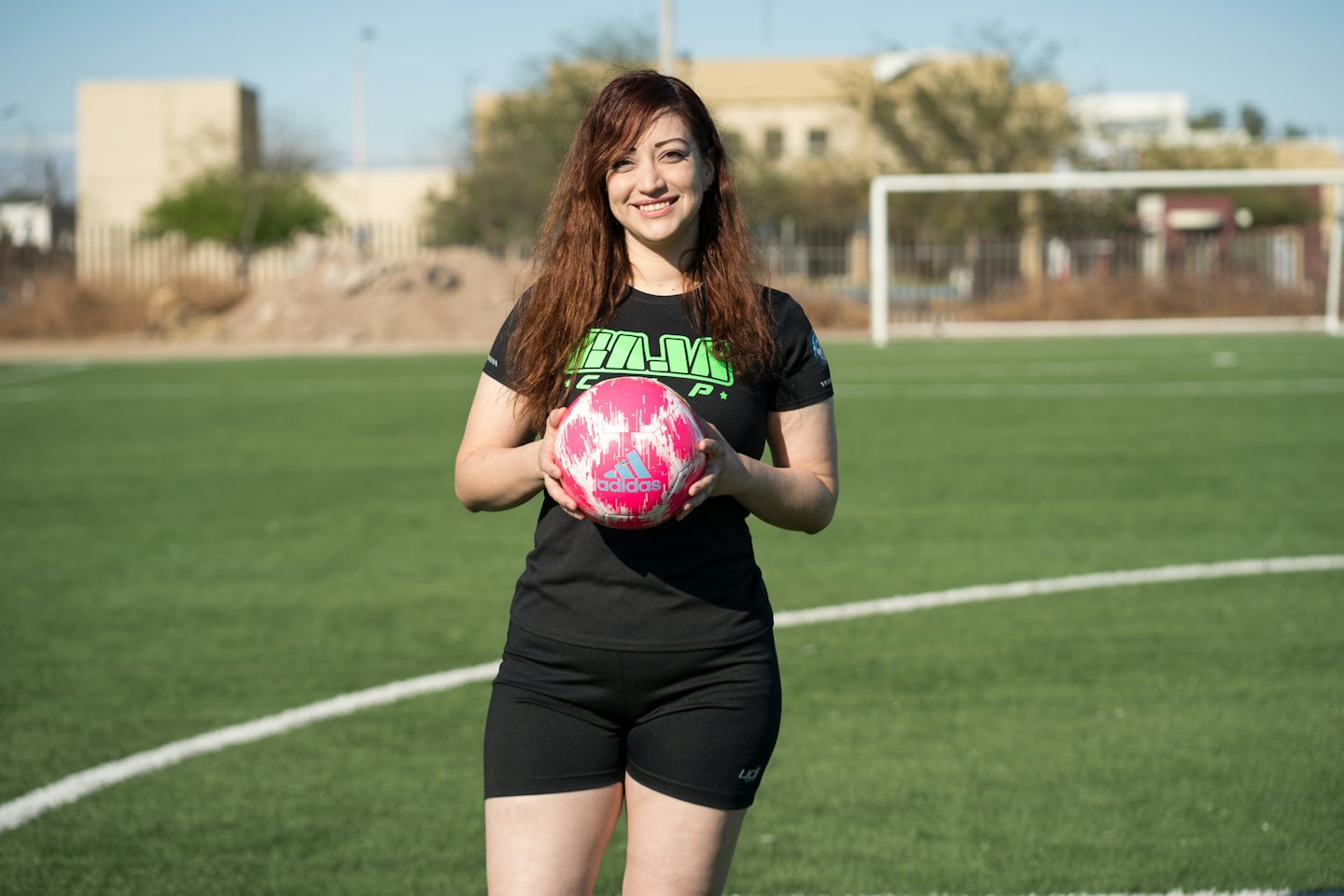 Mujer sonriente con un balón de fútbol en un campo verde.